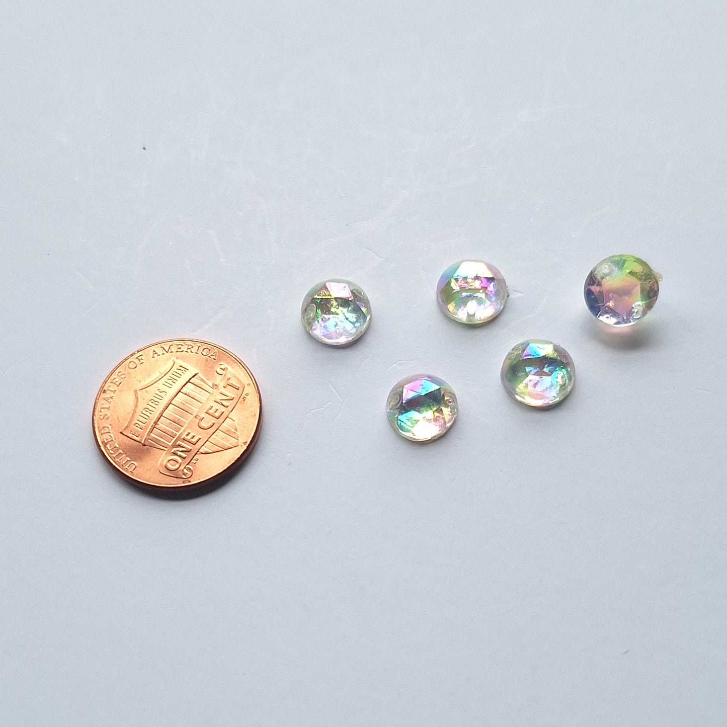 Crystal AB round faceted cabochons arranged in a group of five, with one flipped to show the two-hole sew-on back; penny included for scale.