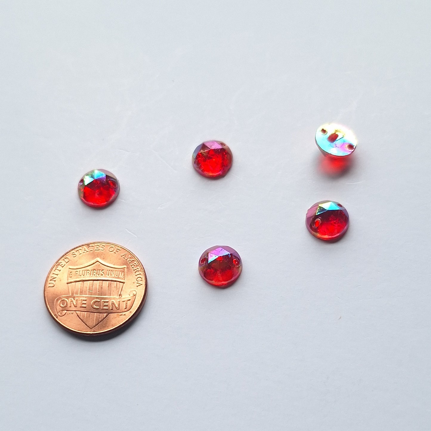 Ruby AB round faceted cabochons arranged in a group of five, with one flipped to show the two-hole sew-on back; penny included for scale.