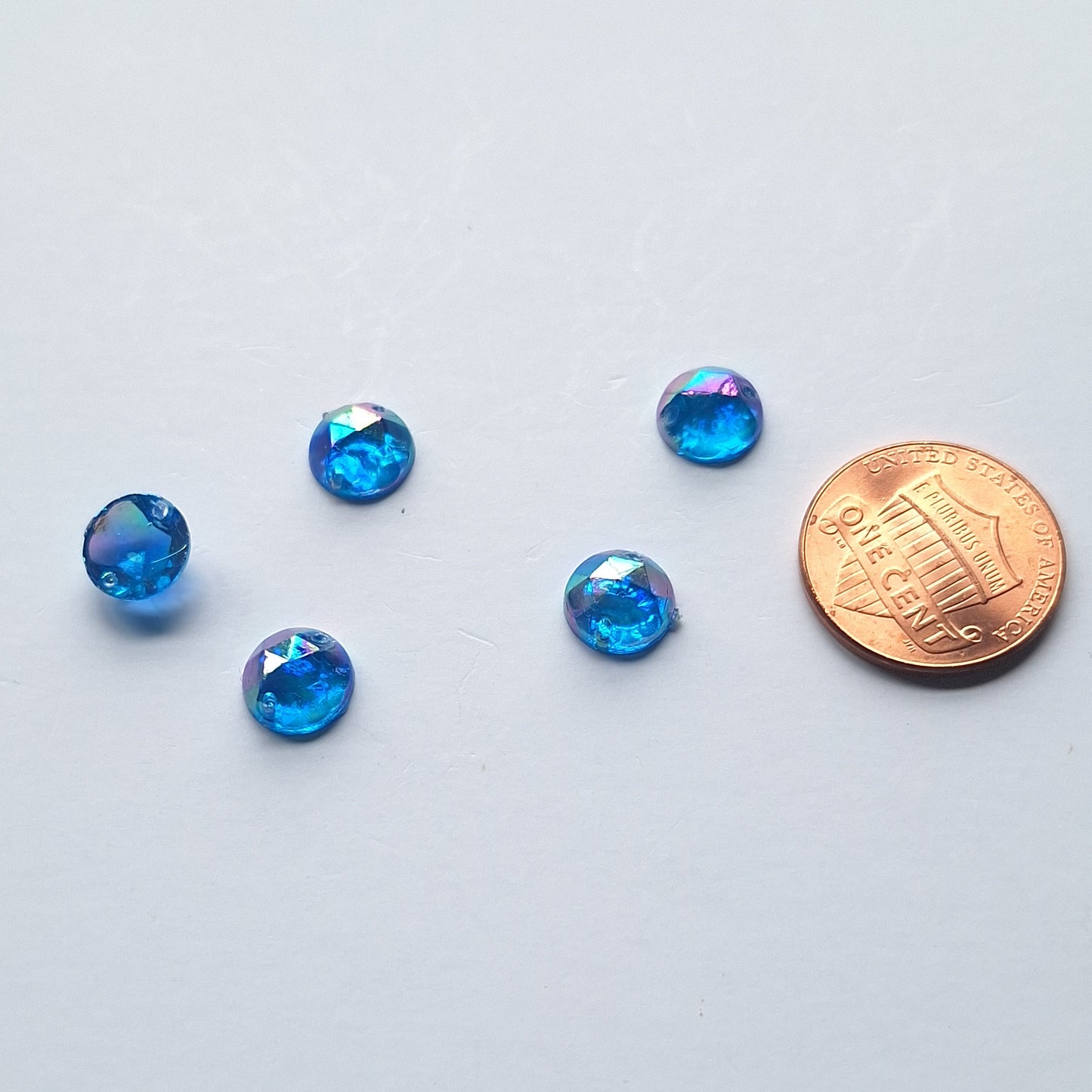 Sapphire AB round faceted cabochons arranged in a group of five, with one flipped to show the two-hole sew-on back; penny included for scale.