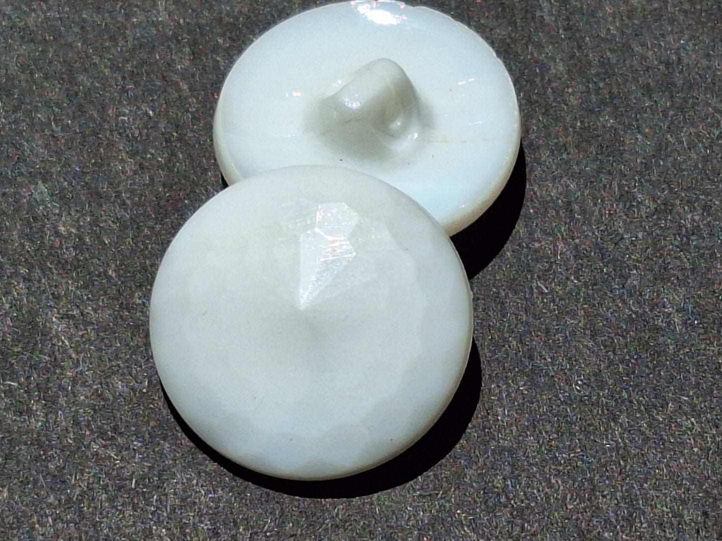 Two white glass shank buttons shown front and back, with a raised multi-facet dome on the face and a smooth molded shank underneath, displayed in direct sunlight to reveal the shallow perimeter facets and deeper central facets.