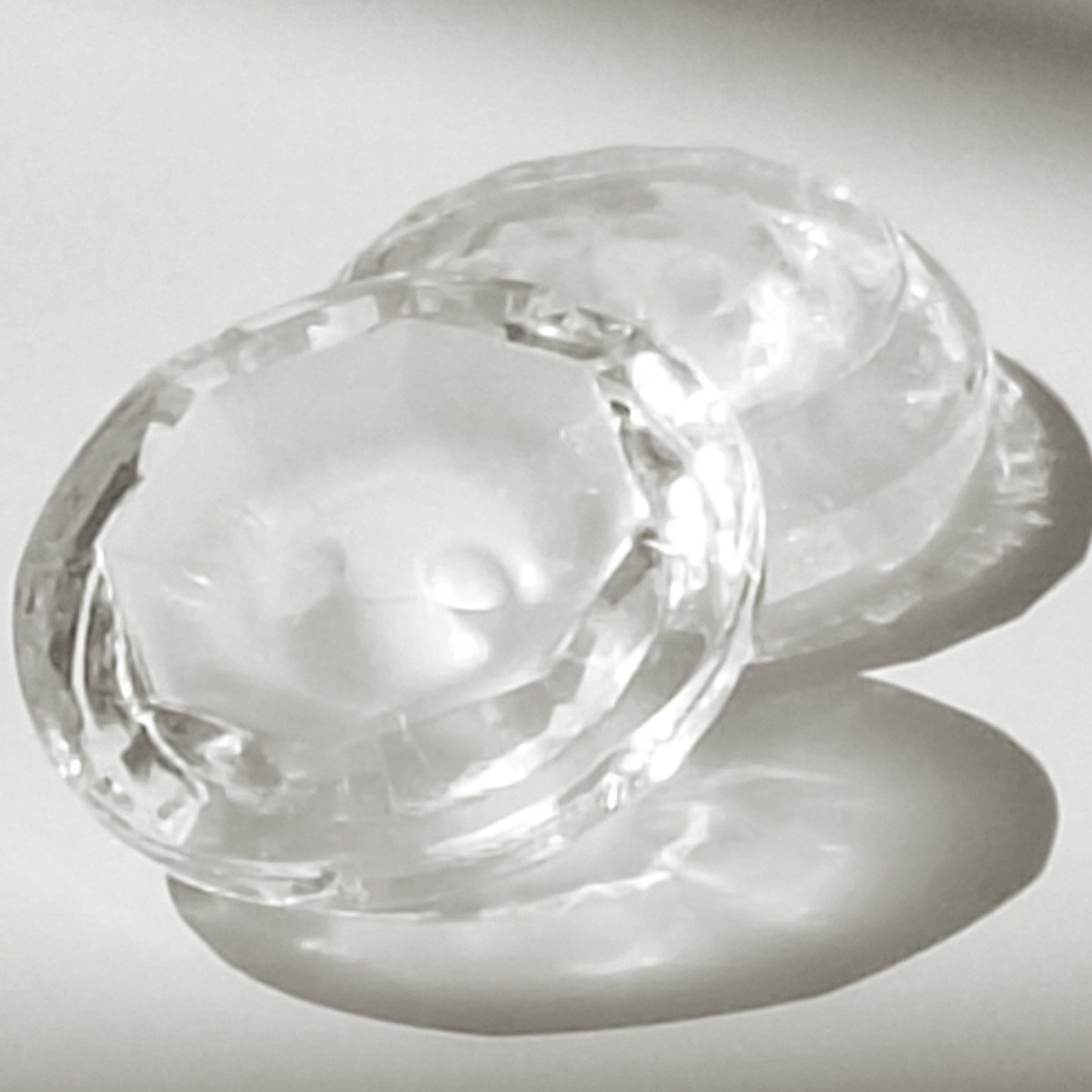 Two clear glass buttons with frosted centers, one facing up and leaning against another facing down, photographed in strong directional light that creates pronounced reflections.